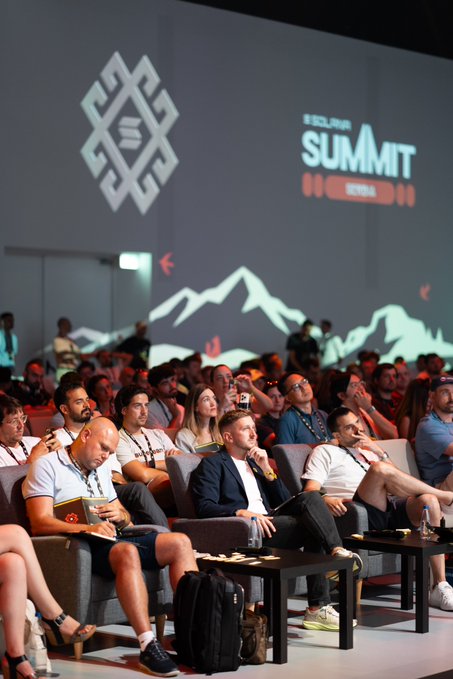 A large audience sits in an auditorium watching a presentation, with a screen in the background displaying a logo and the text 'SUMMIT'.