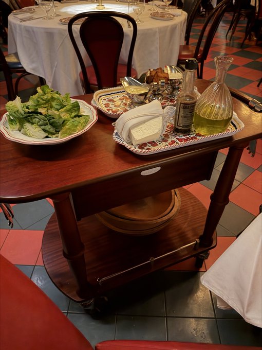 The image shows a wooden serving cart at what appears to be an Italian restaurant, featuring a Caesar salad, bruschetta, a bottle of olive oil, a glass cruet with yellow liquid, and butter on plates. The accompanying post about crypto going parabolic and "AntHood" seems unrelated to the image, as the photo depicts a typical dining scene with no visible connection to cryptocurrency or financial content.