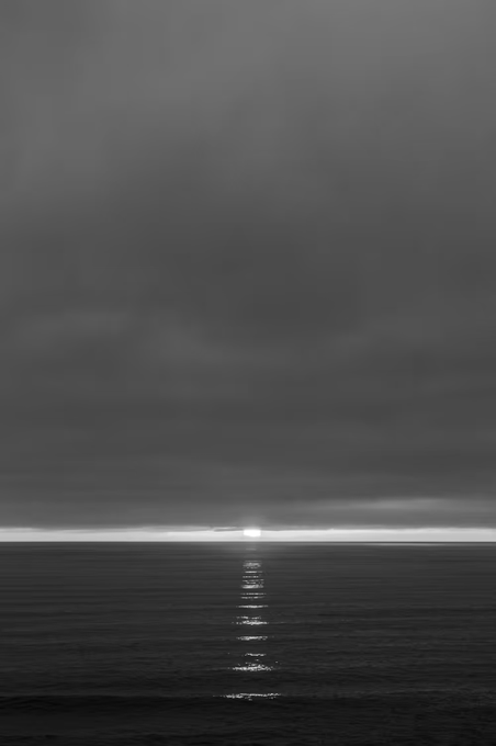 A monochromatic seascape captures a sliver of sunlight breaking through a heavy, overcast sky, casting a shimmering reflection across dark ocean waters. The minimalist, atmospheric composition evokes a contemplative mood fitting for art featured in the Uprising.ART collection shared by Esteban Amaro.