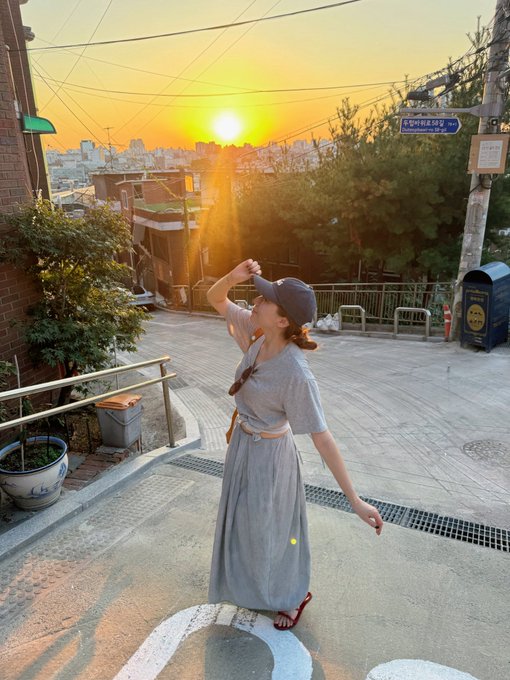 A woman in a long grey dress and dark cap shields her eyes from the bright sunset as she looks out over a hillside neighborhood with a blue street sign.
