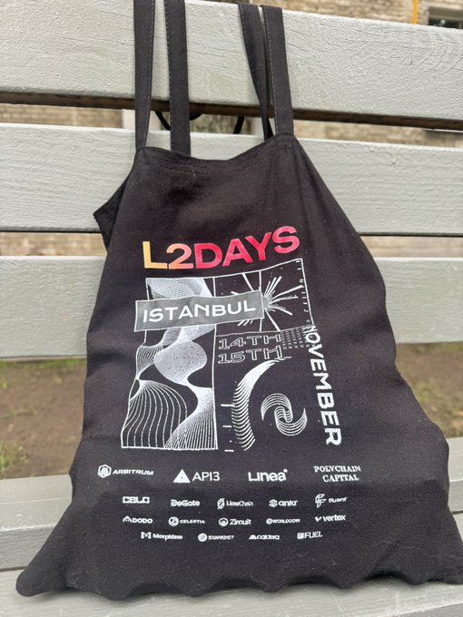 A black tote bag sitting on a wooden bench, displaying 'L2DAYS ISTANBUL 14TH-15TH NOVEMBER' along with sponsor logos including Arbitrum, AP13, Linea, and Polygon Capital.