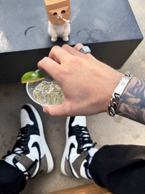 A first-person view shows a person wearing black and white high-top sneakers, holding a clear cocktail with a lime garnish, while a small white dog wearing a paper bag on its head with drawn-on eyes sits nearby.