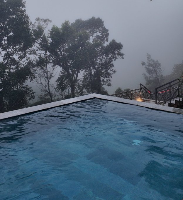 An infinity pool overlooks a misty, fog-covered forest landscape with trees fading into the gray haze, viewed from the pool's edge with metal railings visible on the right.