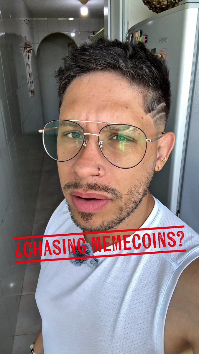 A selfie of a young man with glasses, a nose ring, earrings, and short dark hair, wearing a white shirt in what appears to be a kitchen, with red overlaid text reading '¿CHASING MEMECOINS?'.