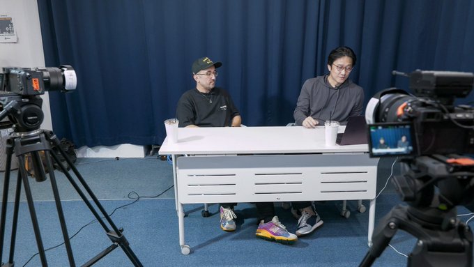Two men sit at a white desk being filmed by professional cameras in what appears to be a podcast or video recording setup.