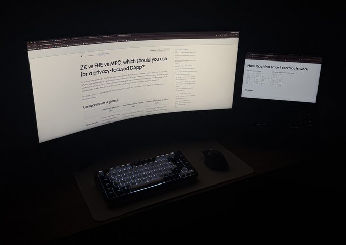 Photo of a dark desk setup featuring a curved monitor displaying an article titled 'ZK vs FHE vs MPC which should you use for a privacy-focused DApp?' alongside a laptop showing 'How Kachina smart contracts work,' a mechanical keyboard, and a mouse.
