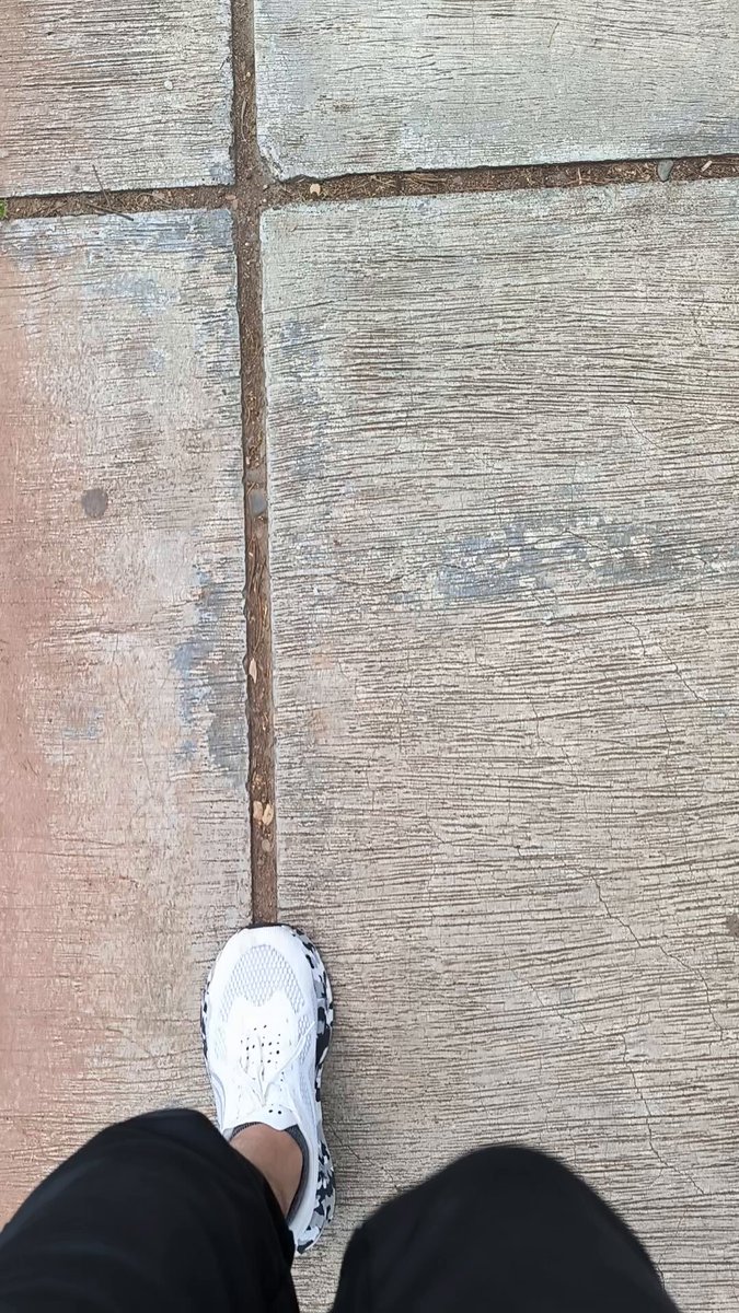 The image shows a person's feet wearing white sneakers, walking along a sidewalk with a concrete surface. The photo appears to be taken from an angle that looks down at the person's feet as they walk. There are no other people or objects visible in the picture.