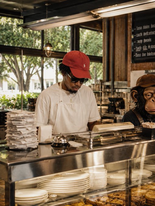The image shows a man wearing a red baseball cap, an apron, and sunglasses standing behind a counter with plates of food displayed in front of him. The counter has several stacks of white plates arranged neatly. There are also some sandwiches on display at the counter. A sign is visible above the counter that reads "gm to everyone I'M finally back and ready to cook. What should I serve you this morning?".