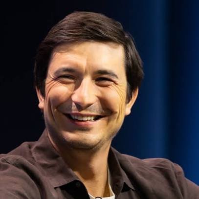 A smiling man with dark hair wearing a brown shirt, photographed against a dark blue background.