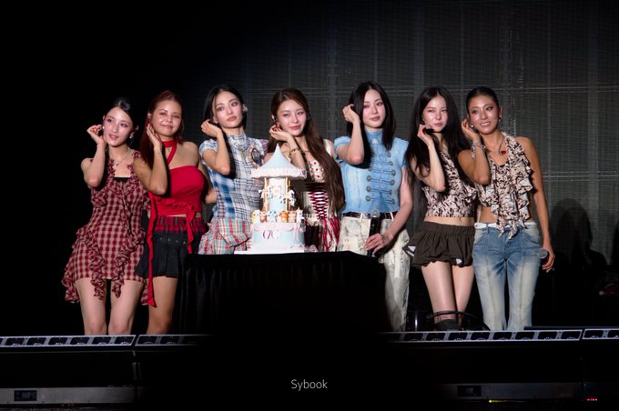 A photograph of seven female K-pop performers from the group TWICE posing together on stage behind a cake at their fan event, credited to 'Sybook'.