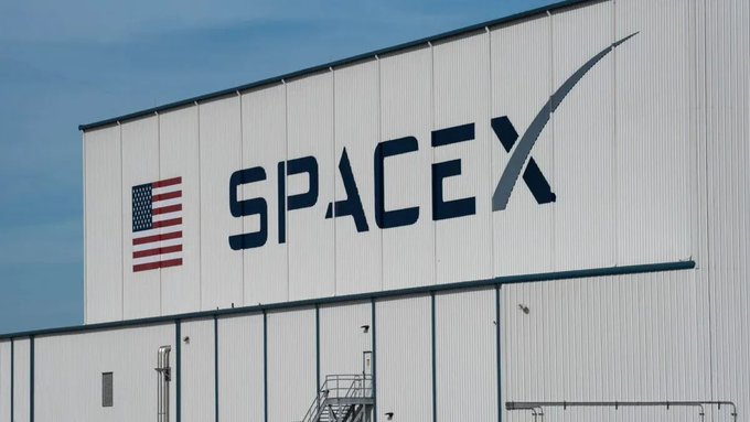 A large white building features the SpaceX logo and an American flag on its exterior wall under a clear blue sky.