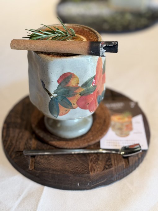 A photograph of a creative cocktail served in a ceramic mug decorated with painted citrus fruits and leaves, garnished with a rosemary sprig and a cinnamon stick, presented on a wooden coaster with a matching spoon.