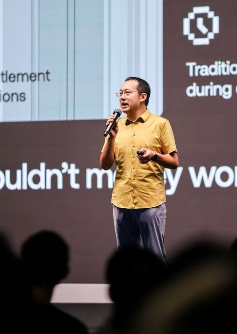 OKX CMO @star_okx speaks on stage at a Singapore event, holding a microphone and presenting in front of a backdrop featuring a clock icon and partial text reading "Tradition during" and "wouldn't m... y wo," while discussing the benefits of their partnership with Mastercard.