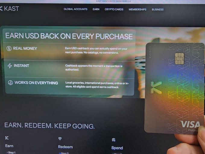 A hand holding a metallic KAST Visa Platinum card in front of a screen displaying their website promoting 'Earn USD back on every purchase' with features for real money, instant cashback, and universal acceptance.