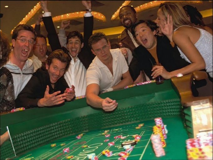 Leonardo DiCaprio and a crowd of others cheer enthusiastically around a green craps table in what appears to be a casino scene from a movie.
