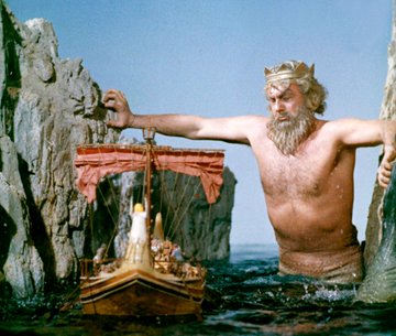 A scene from a vintage fantasy film showing a giant bearded figure reaching toward a small wooden ship with a red sail as it passes between rocky cliffs.