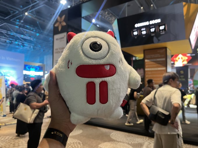A hand holds up a cute plush toy of a white, round, one-eyed monster with red horns and red feet, photographed at what appears to be a convention with booths and people in the background.