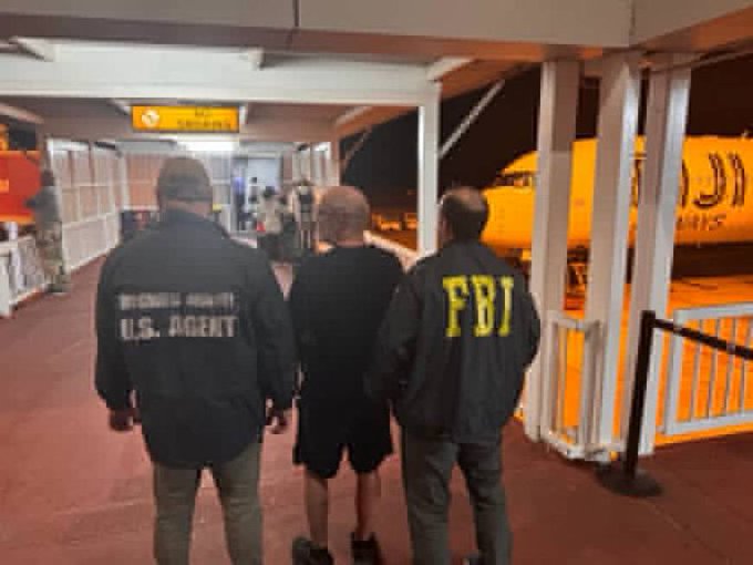 Two federal agents—one wearing a jacket marked "U.S. AGENT" and another with "FBI" emblazoned on his back—escort a handcuffed man in shorts through what appears to be an airport terminal, depicting the extradition of alleged crypto Ponzi schemer Edward Zimbardi from Fiji back to the United States to face charges in his $165 million fraud case.