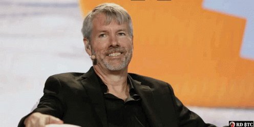 The image features an older man sitting down with his arms crossed, wearing a black suit jacket. He has a big smile on his face as he looks directly into the camera. The background of the image shows a large orange wall that appears to be part of a stage or performance area. There is also text in the image that reads "I would absolutely lose my shit laughing if Saylor managed to trigger a liquidation cascade by selling 32 $BTC and then ATM’d the living hell out of the common to pick up 3,200 BTC at a discount."