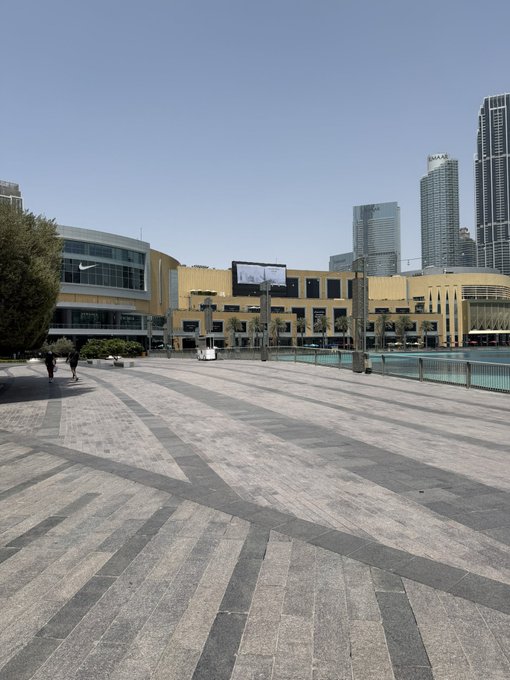 An outdoor plaza with geometric patterned stone tiles leading toward a Nike store and modern buildings, with skyscrapers visible in the background on a clear day.