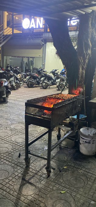 A nighttime street food scene featuring a charcoal grill with meat skewers cooking over glowing coals on a tiled sidewalk, with parked motorbikes and a lit storefront in the background.