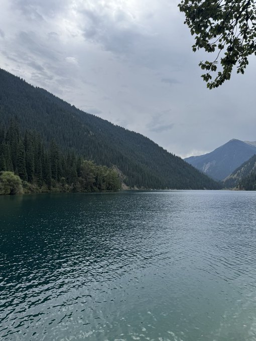 A serene mountain lake with calm teal waters surrounded by forested hills under an overcast sky. The tranquil natural scene perfectly mirrors the post's message of taking a peaceful holiday break to enjoy fresh air while the crypto market surges with XRP, ETH, and BTC all pumping.