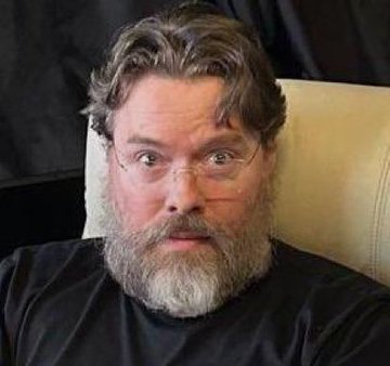 A bearded man with glasses and unkempt hair sits in a leather chair, looking slightly disheveled with an expression of bewildered surprise, resembling the "Gandalf the Great Wizard" look referenced in the post about Richard Heart's detention.