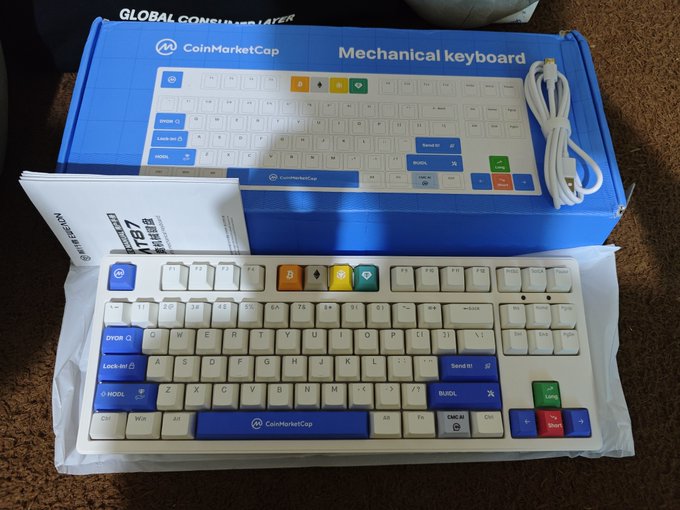 A CoinMarketCap branded mechanical keyboard (NZXT Function TKL) is displayed next to its blue packaging, featuring crypto-themed keycaps with Bitcoin, Ethereum, and BNB symbols, as well as trading labels like "LONG," "BUY," "SELL," and "Send it!"—a perfect prize for upgrading a trader's workstation.