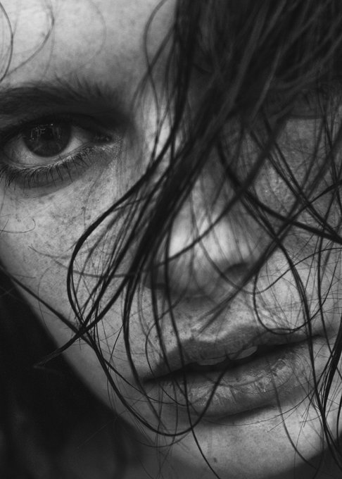 A high-contrast, black and white close-up portrait of a person's face, with strands of dark hair partially obscuring their features and focusing on their intense gaze.