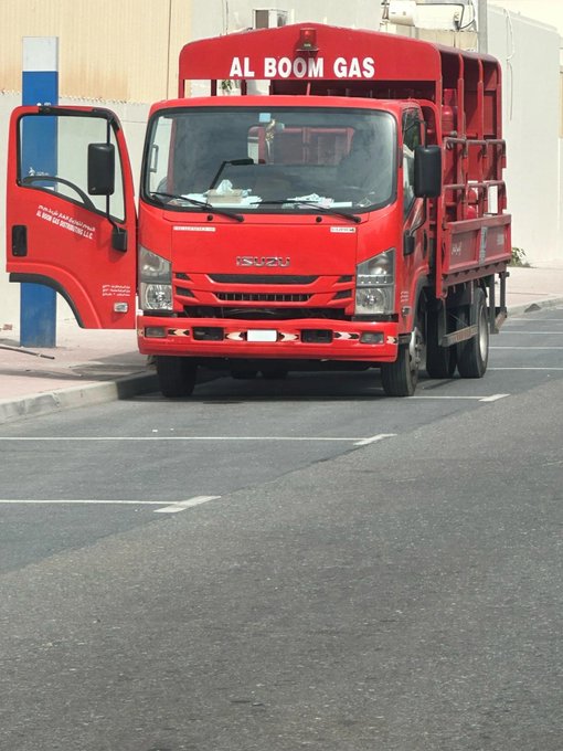 A red Isuzu truck labeled "AL BOOM GAS" is parked on a street with its driver's side door open.
