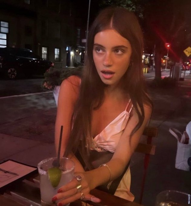 A young woman with long brown hair, wearing a pink tie-front top, sits at an outdoor nighttime café table with a lime-garnished cocktail.