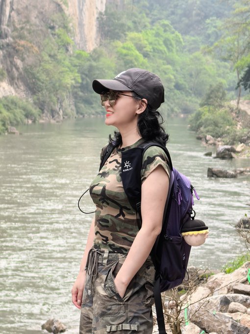 A woman in a camouflage outfit and cap stands by a scenic river, wearing a backpack with a plush keychain, exuding a confident, carefree spirit that matches the post's message of self-assurance and "charging ahead"—with the hashtag #BTC and #Crypto tying the adventurous independence to the bold financial spirit of going your own way.