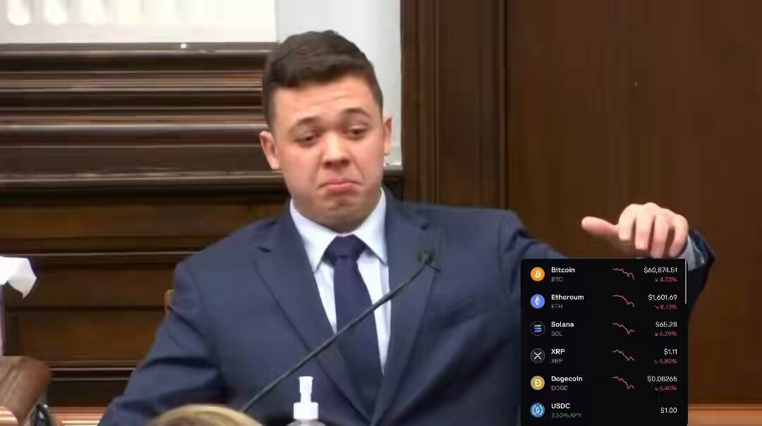 The image shows a man wearing a suit standing at a podium with a microphone in front of him. He is pointing towards a computer screen displaying a list of cryptocurrency names, including Bitcoin, Etherium, Ripple, Litecoin, and others. The man appears to be giving a speech or presentation about the various cryptocurrencies on display.