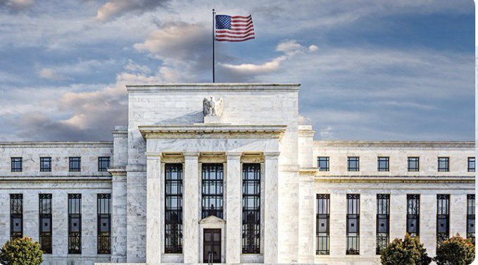 The image features a large white building with an American flag flying atop of it. The building appears to be a government or financial institution, as indicated by its architectural style and design elements such as columns and arches. The presence of the American flag suggests that the building may be associated with the Federal Reserve Bank of the United States (Fed).