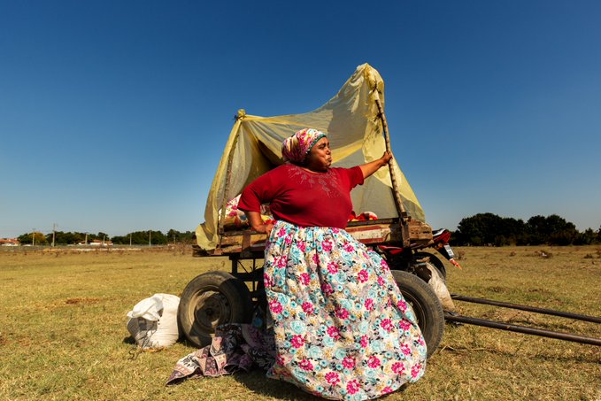 A woman in traditional clothing—a red top, floral skirt, and headscarf—posing beside a wooden cart covered with a yellow tarp in an open grassy field under a clear blue sky.