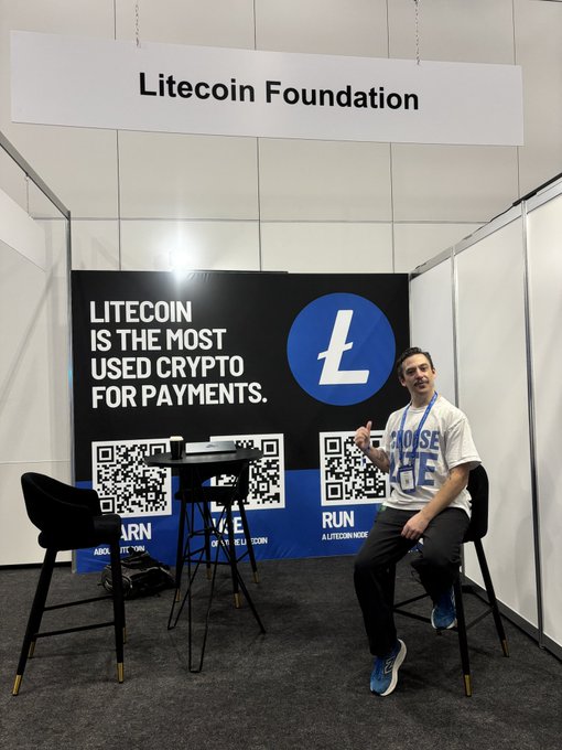 A young man wearing a white shirt sits at an information desk with two black stools behind him while he poses for a picture. The backdrop displays a banner that reads "Litecoin running full speed in New Zealand". Additionally, there are three tables visible in the image and a chair positioned near one of them.