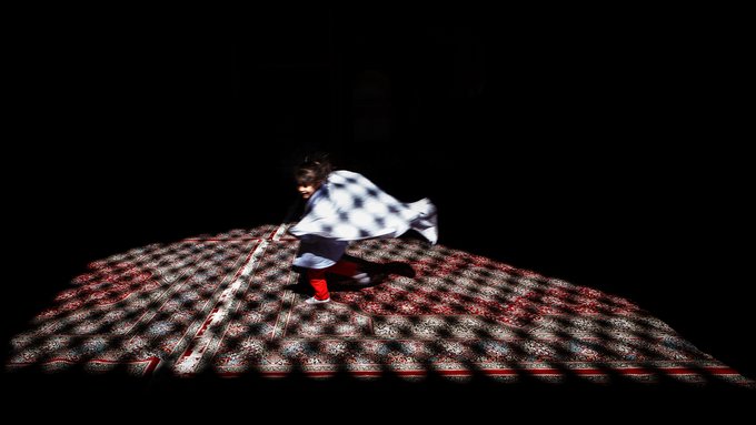 A performer in a white and blue checkered outfit with red boots dancing on a stage with a patterned, illuminated floor in a dark setting.