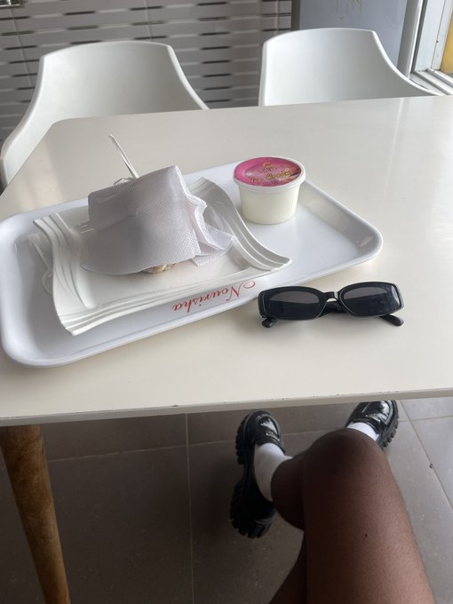 A first-person view of someone sitting at a white table with a tray holding a wrapped burger, a cup of pudding or yogurt, black sunglasses, and a napkin, with their legs and black platform shoes visible beneath the table.