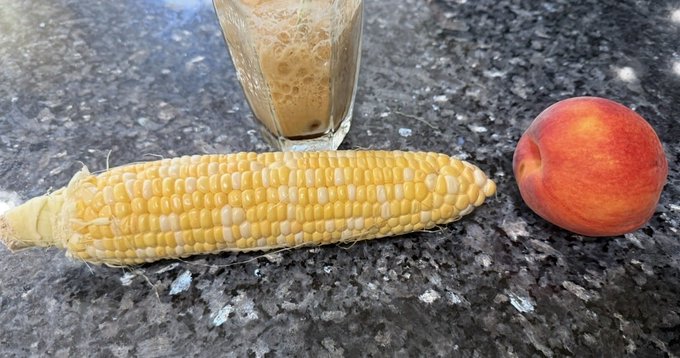 A yellow ear of corn, a peach, and a glass of a foamy beverage arranged on a dark granite countertop.