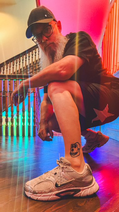 A man with a long white beard, glasses, and a cap crouches on a wooden staircase lit with rainbow colors, wearing Nike sneakers and shorts with a star.