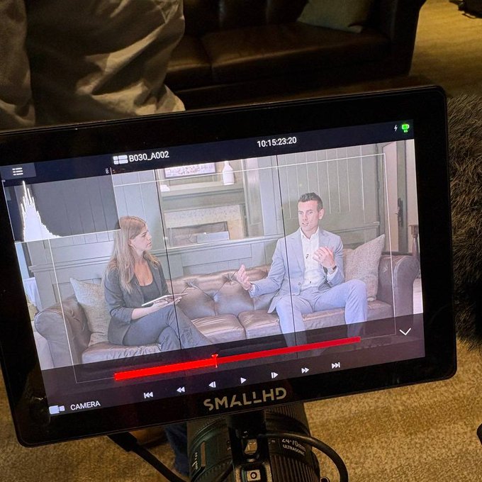 A SmallHD camera monitor displays a video feed of a man and woman in an interview setting on a leather couch, with a timestamp reading 10:15:23:20 and the camera label B030_A002.