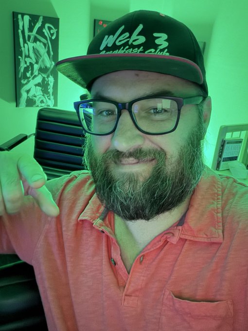A bearded man wearing glasses and a black 'Web 3 Breakfast Club' cap smiles and points at the camera, lit with a green hue in what appears to be an office setting.