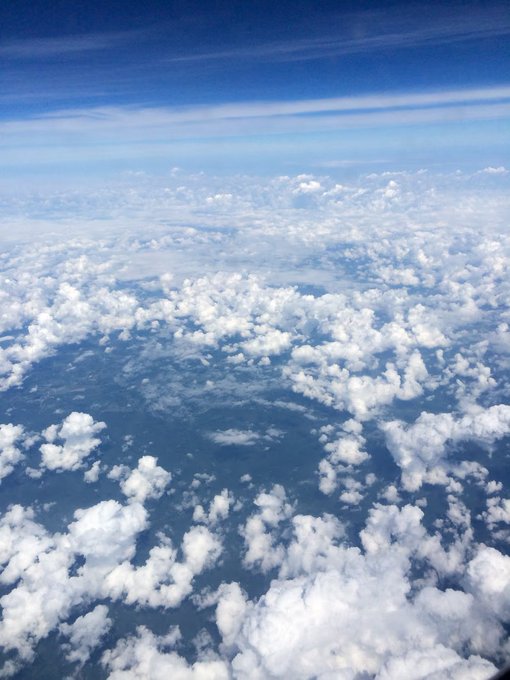An aerial view from an airplane window shows fluffy white clouds scattered across a landscape with visible green and brown terrain below, stretching to the horizon under a blue sky with high cirrus clouds.