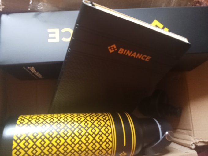 Binance-branded merchandise including a notebook, packaging box, and a yellow and black tumbler water bottle in a cardboard shipping box.