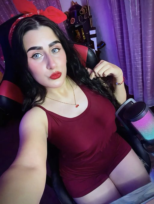 A woman with dark hair and bold makeup sits in a gaming chair wearing a maroon dress and a red bow headband, with a colorful RGB-lit device next to her.