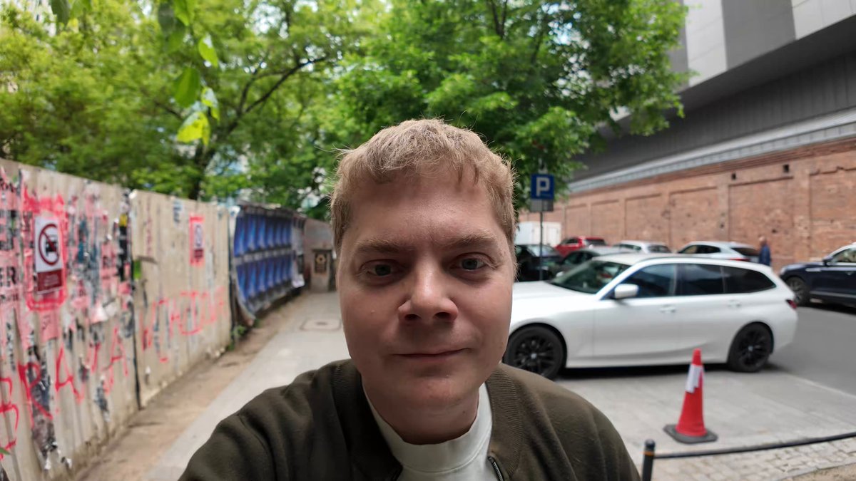 The image shows a man wearing a green jacket standing next to a brick building with cars parked nearby. The man appears to be posing for a photo, possibly for a marketing campaign or promotional material related to the building or the vehicles present. The focus of the image is on the man and his surroundings, highlighting the urban setting and the connection between the man's presence in the scene and the content of the post.