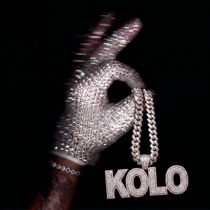 The image features a hand holding up a silver necklace that has the word "KOLO" written on it, along with the phrase "Good morning, ct Isn' t it funny how many people buy crypto but still treat it like something they can't actually use day to day". The person is wearing a glove and there are diamonds visible in the background.
