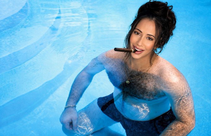 A topless woman with dark hair smiles while holding a cigar in her mouth as she poses in a swimming pool. The image's playful, defiant tone parallels the post's message of perseverance and resilience, reinforcing a "never again" attitude with bold confidence.
