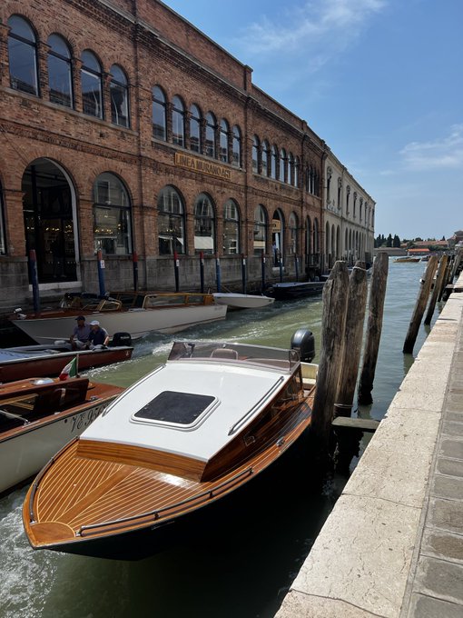 A scenic photograph of wooden boats moored in a canal alongside a historic brick building in Venice, with one boat displaying 'LANCIA E MOBILIO ARTI' signage on the building.