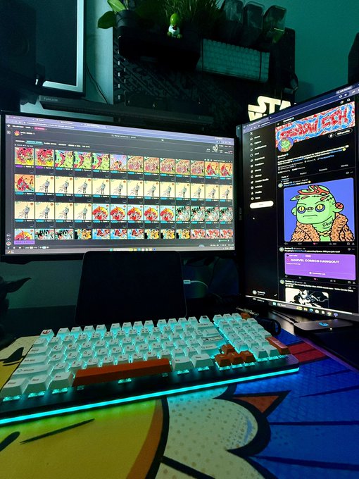 A desk setup with a mechanical keyboard featuring teal backlighting, dual monitors displaying an NFT collection gallery and YouTube page with a cartoon character.